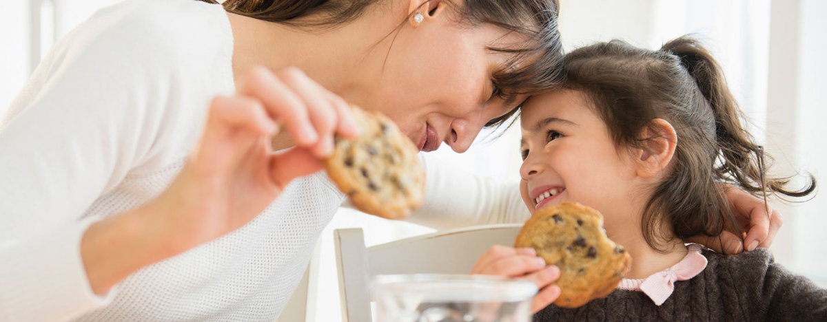 A mom and daughter smiling while eating chocolate chip cookies